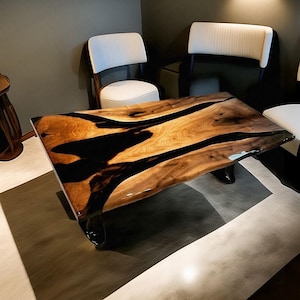 Op de afbeelding: Een rechthoekige houten salontafel met een glanzende afwerking. De tafel heeft een natuurlijk houtnerfpatroon met donkere zwarte harsinlays. De tafel wordt ondersteund door zwarte, gebogen poten. Witte stoelen omringen de tafel.