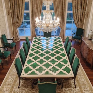 Puede incluir: Un comedor formal con una gran mesa rectangular con un patrón geométrico verde y blanco. La mesa está rodeada de sillas tapizadas verdes. Un candelabro de cristal cuelga encima, y la habitación tiene grandes ventanas con cortinas doradas.