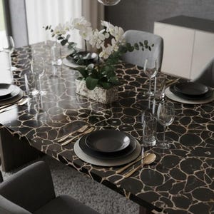 May include: A dining table set for a formal dinner. The table has a dark, patterned surface with black plates, gold silverware, and clear glassware. Gray chairs surround the table, and a floral arrangement sits in the center.