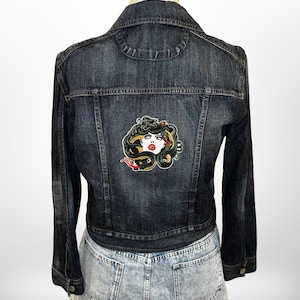 Könnte beinhalten: Dunkelblaue Jeansjacke mit einem Medusa-Aufnäher auf dem Rücken. Der Aufnäher zeigt eine Frau mit Schlangen als Haare, roten Lippen und einer roten Zunge. Die Jacke wird über einem hellblauen Jeansrock getragen.