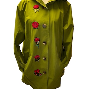 Może przedstawiać: Oliwkowy płaszcz z kapturem z czterema aplikacjami w kształcie czerwonych róż. Płaszcz ma zapięcie na guziki, dwie kieszenie i kaptur z podszewką w kratę. Płaszcz jest prezentowany na manekinie.