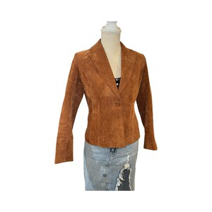 May include: A brown suede blazer with a notched lapel and a single button closure. The jacket has long sleeves and a tailored fit. The blazer is displayed on a mannequin.