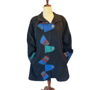 Op de afbeelding: Zwarte wollen jas met kleurrijke geometrische patches en knevelsluitingen. De jas heeft blauwe, groene en rode accenten, met een klassieke kraag en zakken. De jas wordt gepresenteerd op een mannequin.