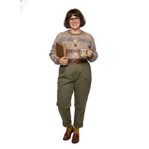 May include: A person wearing glasses, a patterned sweater, olive green pants, and brown loafers. They hold a book and a mug with a floral design. The outfit includes a brown belt and gold necklaces.