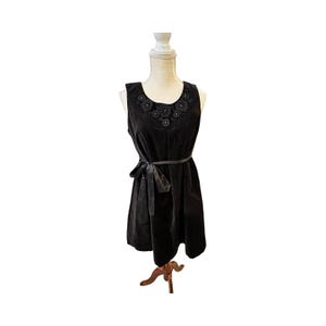 May include: A sleeveless black dress with a round neckline adorned with floral embellishments. The dress features a fitted bodice, a flared skirt, and a black ribbon belt tied at the waist. The dress is displayed on a mannequin.