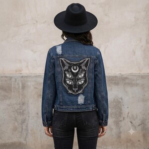 Könnte beinhalten: Eine dunkelblaue Jeansjacke mit einem großen schwarz-weißen Aufnäher auf dem Rücken, der eine Katze mit vier Augen und einem Mondsichelsymbol auf der Stirn zeigt. Die Jacke hat abgenutzte Details und wird mit einem schwarzen Hut getragen.