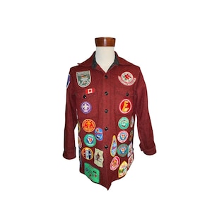 Puede incluir: Una camisa de lana granate con numerosos parches coloridos. La camisa tiene cuello y cierre de botones. Los parches incluyen varias formas y diseños, con texto y símbolos. La camisa se exhibe en un maniquí.