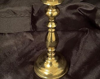May include: A brass candlestick holder with a round base and a slender, ornate stem. The top features a circular cup to hold a candle. The polished brass reflects light, creating a warm, inviting glow. A decorative accent for any home.