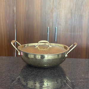 Può includere: Un wok dorato martellato con coperchio in vetro e due manici laterali. Il wok ha una forma arrotondata e una superficie strutturata. Il coperchio ha una piccola maniglia. Il wok è appoggiato su un piano scuro.