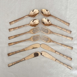 Könnte beinhalten: Ein Satz goldfarbenes Besteck, einschließlich Löffel, Gabeln und Buttermessern. Die Griffe haben ein bambusartiges Design. Die Anordnung der Utensilien ist symmetrisch, mit den Löffeln und Messern oben und unten und den Gabeln in der Mitte.