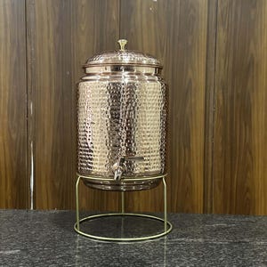 May include: A copper-colored beverage dispenser with a hammered texture, a spigot, and a decorative lid. It sits on a gold-colored metal stand. The dispenser is designed for serving drinks and adds a touch of elegance to any event.