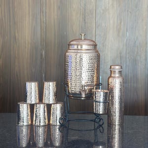 May include: A copper-colored water dispenser set with a hammered texture. The set includes a large dispenser with a spigot, a water bottle, and several small drinking glasses. The dispenser sits on a black metal stand.