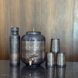 Può includere: Un set di erogatore d'acqua grigio scuro con una bottiglia abbinata e quattro tazze. Il set presenta un ornato motivo floreale dorato. L'erogatore ha un rubinetto e le tazze sono cilindriche. Il set è esposto su una superficie scura.