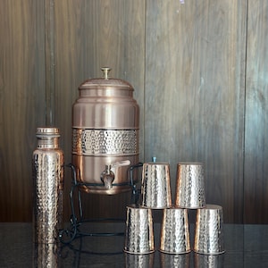 May include: A copper-colored water dispenser set with a hammered texture. The set includes a tall water bottle, a large dispenser with a spigot, and six matching cups. The dispenser sits on a black metal stand. The background is a wooden wall.