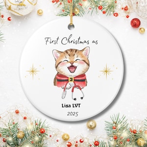 Könnte beinhalten: Runder Keramik-Weihnachtsschmuck mit einem lächelnden Tabby-Kätzchen in einem Weihnachtsmann-Outfit, das ein Stethoskop hält. Der Text lautet "First Christmas as" über der Katze, mit "Lisa LVT 2025" darunter. Der Schmuck hat eine goldene Schnur zum Aufhängen.