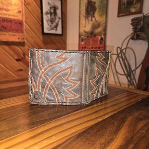 May include: A brown leather wallet with a western-style design. The wallet features a stitched pattern in shades of orange and cream, resembling a stylized leaf or floral motif. The wallet is rectangular and appears to be folded.