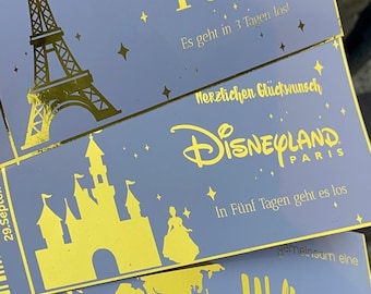 May include: Three rectangular cards with a blue background and gold foil accents. The top card features the Eiffel Tower and text reading Wir machen eine Reise nach Paris. The middle card shows a castle and Disneyland Paris logo. The bottom card displays a world map.