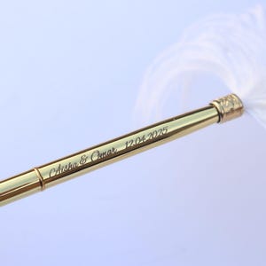 May include: A gold-toned metal quill pen featuring a white feather plume. The barrel of the pen is engraved with the text Aisha & Omar 12.04.2025 in a cursive script font. The pen has a smooth, reflective surface and a metallic finish.