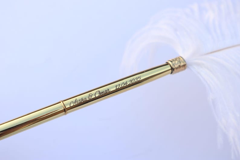 May include: A gold-toned metal quill pen featuring a white feather plume. The barrel of the pen is engraved with the text Aisha & Omar 12.04.2025 in a cursive script font. The pen has a smooth, reflective surface and a metallic finish.