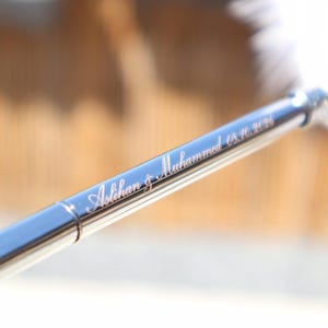 May include: A sleek, metallic pen with a polished finish featuring the engraved names Aslihan & Muhammed and the date 8.16.2026 in a cursive script. The writing instrument has a slender, cylindrical body with a reflective surface.