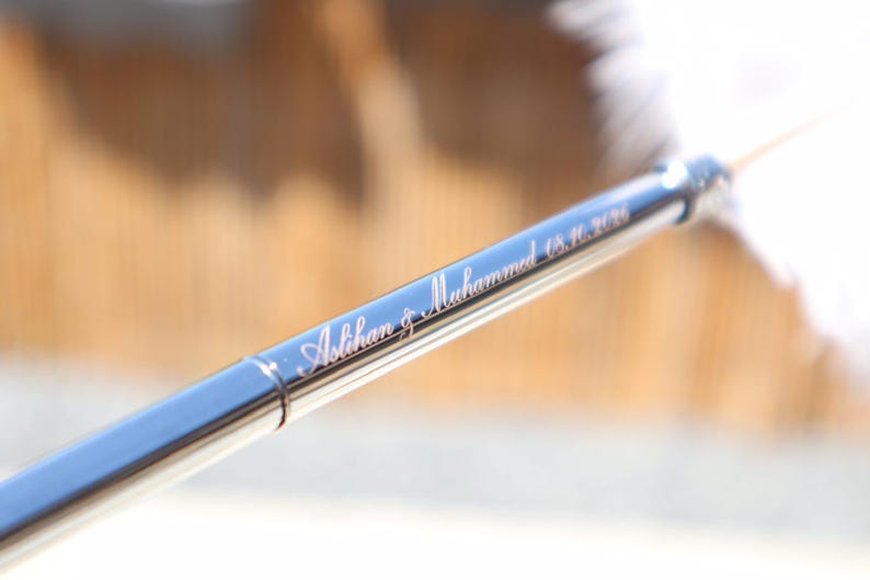 May include: A sleek, metallic pen with a polished finish featuring the engraved names Aslihan & Muhammed and the date 8.16.2026 in a cursive script. The writing instrument has a slender, cylindrical body with a reflective surface.