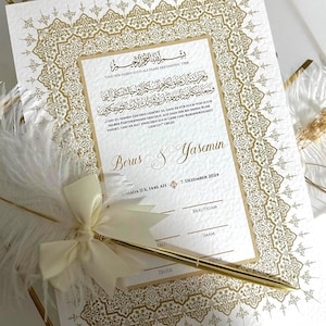 Könnte beinhalten: Eine weiße Hochzeitsurkunde mit einem aufwendigen Goldfolienrand, arabischem und deutschem Text sowie den Namen Berus & Yasemin. Das Dokument enthält Zeilen für Unterschriften und einen goldenen Stift, der mit einer weißen Feder verziert ist.
