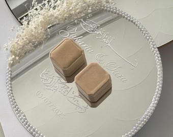 May include: A round, mirrored tray with a pearl-beaded edge, holding two beige ring boxes. The tray is engraved with Arabic script and the names "Zakaria & Sara" along with the date "05/07/2025". White dried flowers adorn the top.