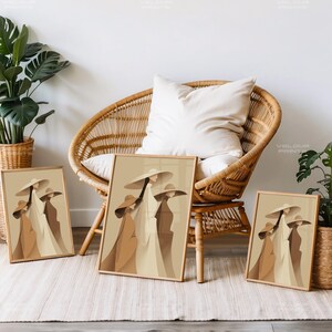 May include: Three framed art prints featuring a minimalist illustration of figures in wide-brimmed hats and long coats. The artwork uses a palette of beige, brown, and cream tones. The prints are displayed near a wicker chair and potted plants.