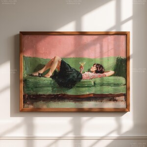 May include: A framed art print depicts a person reclining on a green sofa, holding a cocktail. The artwork features a pink background and a wooden frame. The person is wearing a dark skirt and a pink and white patterned top.