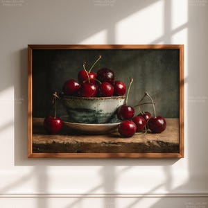 May include: A framed still life print featuring a bowl of ripe cherries. The cherries are a deep red color and are arranged in a ceramic bowl with a blue and white design. The print is set on a wooden surface.