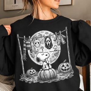 May include: A black crewneck sweatshirt featuring a white graphic print of Snoopy sitting on a pumpkin under a full moon. A clothesline above displays a striped sweater, a hockey mask, a ghost mask, and overalls, with Woodstock nearby.
