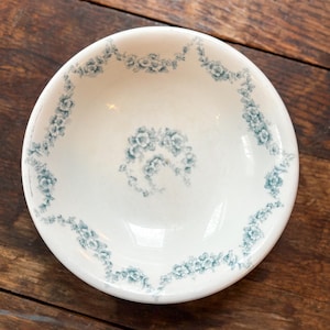 May include: A white ceramic bowl with a delicate blue floral pattern around the rim and center. The bowl is round and sits on a wooden surface. The floral design is a repeating pattern of small flowers and leaves.