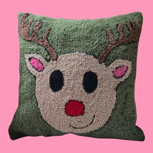 Peut inclure: Un coussin carré vert avec un motif de renne. Le renne a des bois bruns, des yeux noirs, un nez rouge et des oreilles roses. Le coussin est fait d'un matériau texturé, parfait pour la décoration de Noël.