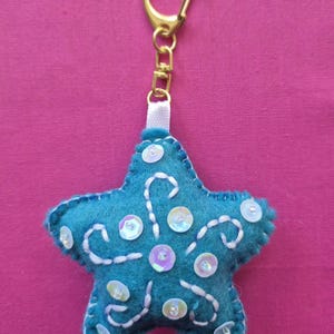 以下が含まれることがあります： 白の刺繍と虹色のスパンコールが施された、ターコイズ色の星型キーホルダー。 キーホルダーには金色の留め金と白いリボンループが付いています。 背景は無地のピンク色です。