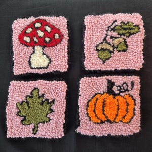Op de afbeelding: Set van vier vierkante, handgetufte onderzetters. Elke onderzetter heeft een ander ontwerp: een rode paddenstoel met witte stippen, groene bladeren, een groen blad en een oranje pompoen. De onderzetters hebben een roze achtergrond en zwarte randen.
