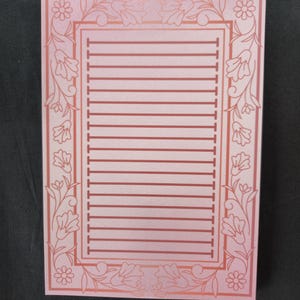Op de afbeelding: Een roze notitieblok met een bloemenrand en gelinieerde pagina's. De rand heeft een herhalend patroon van bloemen en bladeren in een donkerdere roze tint. De gelinieerde pagina's zijn een lichtere roze tint.