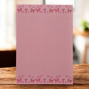 Op de afbeelding: Een roze notitieblok met een rand van aquarel strikken in roze en perziktinten. Het rechthoekige notitieblok ligt op een houten oppervlak. De achtergrond is wazig, wat duidt op een natuurlijke omgeving.