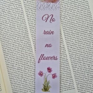 Op de afbeelding: Bladwijzer met de tekst "No rain no flowers" in sierlijke letters, met een aquarel van een wolk en regendruppels en roze bloemen. De bladwijzer ligt op een open boek.