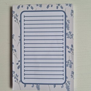 Op de afbeelding: Een rechthoekig notitieblok met een lichtgrijze achtergrond en een blauw bloemenpatroon. Het notitieblok heeft een blauwe rand en horizontale blauwe lijnen om op te schrijven. Het notitieblok is ontworpen om notities te maken.