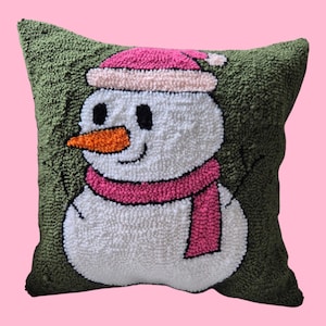 Pode incluir: Uma almofada quadrada com um desenho de boneco de neve. O boneco de neve tem um corpo branco, um nariz de cenoura laranja e um cachecol e chapéu rosa. O fundo é verde. Decoração festiva de inverno.