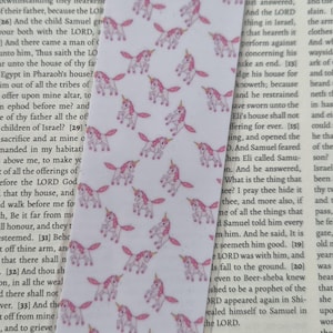 Op de afbeelding: Een witte bladwijzer met een herhaald patroon van roze eenhoorns. De bladwijzer is geplaatst op een pagina van een boek met tekst.