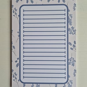 Op de afbeelding: Een rechthoekig notitieblok met een lichtroze achtergrond en een blauw bloemenpatroon. Het notitieblok heeft een blauwe rand en horizontale blauwe lijnen om op te schrijven. Het ontwerp is eenvoudig en elegant.