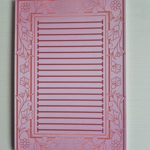 Op de afbeelding: Een roze gelijnd notitieblok met een decoratieve bloemenrand. Het notitieblok heeft een rechthoekig frame met een reeks horizontale lijnen om op te schrijven. De rand heeft gestileerde bloemen en bladeren in een donkerdere roze tint.