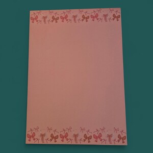 Op de afbeelding: Een vel roze briefpapier met een rand van aquarel strikken in roze en rode tinten. De strikken zijn gelijkmatig verdeeld langs de boven- en onderranden van het papier. De achtergrond is effen roze.