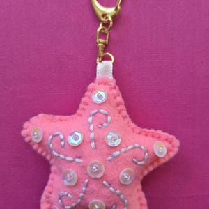 Puede incluir: Llavero en forma de estrella de fieltro rosa con lentejuelas blancas y detalles bordados en plata. El llavero tiene un cierre dorado y un lazo de cinta blanca. El fondo es de color rosa liso.