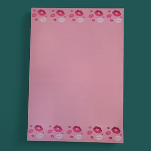 Op de afbeelding: Een roze notitieblok met een rand van rode, roze en witte lippenillustraties. Het rechthoekige notitieblok heeft een effen roze achtergrond, met de lippenontwerpen langs de boven- en onderranden.