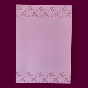 Op de afbeelding: Een rechthoekig, lichtroze notitieblok met een rand van omrande harten in roze en rode tinten. De harten zijn in een herhalend patroon langs de boven- en onderranden gerangschikt, waardoor een decoratief kader ontstaat.