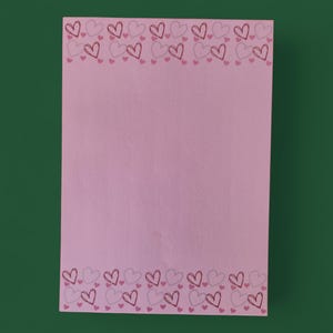 Op de afbeelding: Een roze notitieblok met een rand van omrande harten in roze en grijstinten. De harten zijn gerangschikt in een herhalend patroon langs de boven- en onderranden. De achtergrond is effen roze.