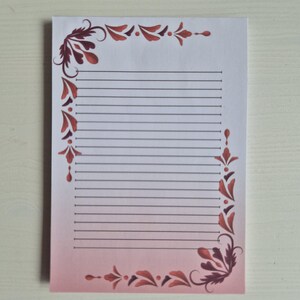 Op de afbeelding: Een gelijnd notitieblok met een decoratieve rand. De rand heeft een kastanjebruin bloemmotief op een roze gradiënt achtergrond. Het notitieblok heeft horizontale lijnen om op te schrijven. Het papier is wit.