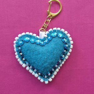 Puede incluir: Un llavero en forma de corazón turquesa con un cierre dorado. El corazón está decorado con lentejuelas azules y ribete de cuentas blancas. El fondo es de color rosa liso.